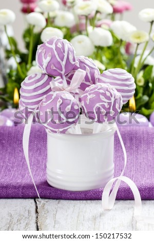 Lilac cake pops in white ceramic jar. White and pink daisies in the background. Party table setting.