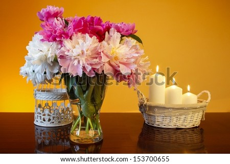 Bouquet of peonies in glass vase and basket of candles on wooden table, on orange background