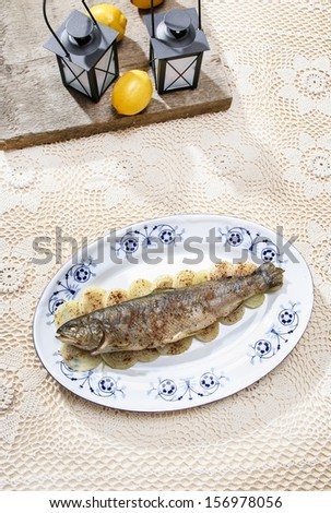 Fish plate in greek tavern. Roasted trout on onion. Pretty beige handmade table cloth. Decorative lanterns and lemons.