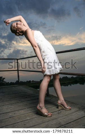 Beautiful young woman in a white summer dress dancing at the beach party. Pretty girl on wooden pier at sunset.