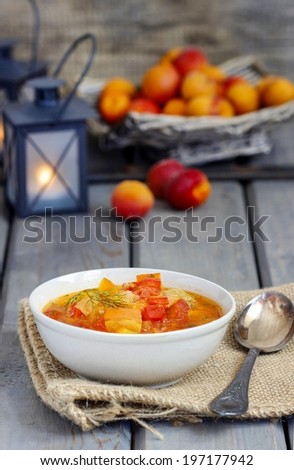 Red vegetable soup in ceramic plate on a rustic wooden table. Lanterns and fruits in the background.