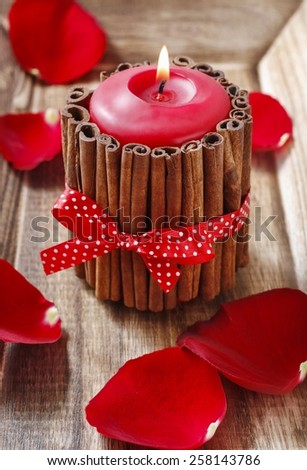 Red scented candle decorated with cinnamon sticks. Rose petals around.