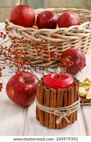 Candle decorated with cinnamon sticks and basket of apples in the background.