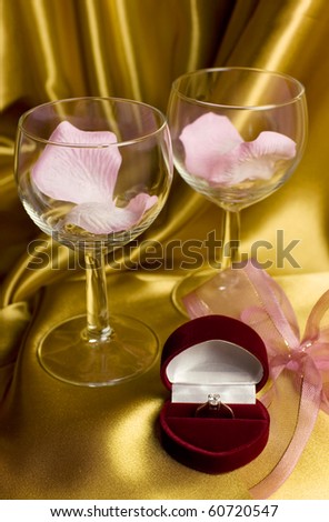 A diamond engagement ring in a heart-shaped dark red velvet box with two glassses and pink flower petals - golden satin background.
