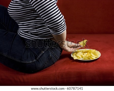 A fat woman sitting on a sofa with chips on a plate, grabing chips behind her back - cheating and failing in her diet.