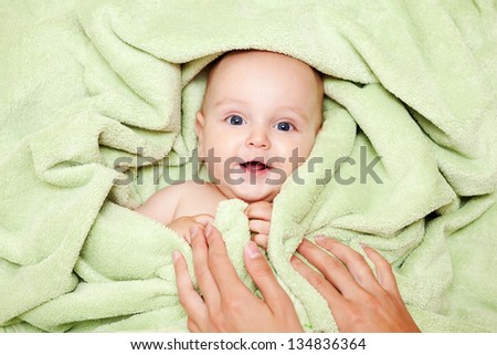 Caucasian baby boy covered with green towel joyfully smiles at camera with mother\'s hands on him