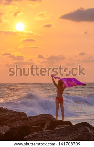 Silhouette of woman that stands against sunset sun at the seashore and waves with her pareo