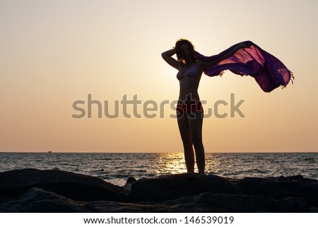 Silhouette of woman that stands against sunset sun at the seashore and waves with her pareo
