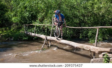 Man carries his bike by log over dark river