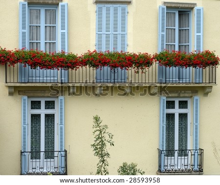 Balcony with red flowers on old house, blue windows and yellow wall