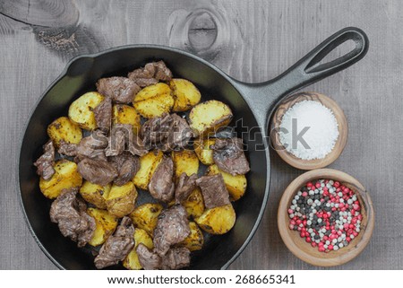 Roast meat with potato in a cast iron skillet and two wooden bowls with salt and multicolored pepper on a wooden table
