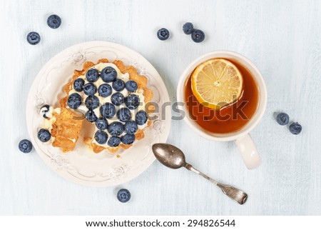 Sweet cake with blueberries, butter cream and syrup , as well as a cup of black tea with a slice of lemon, top view