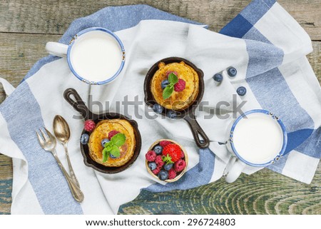 Pancakes in cast-iron frying pan with fresh berries on the background of the old wooden boards and two cups of milk