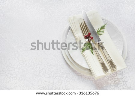 Silver knife and fork, and red holly berries and green thuja branches lie on the white porcelain plate, which is located on a table covered with a white tablecloth, top view