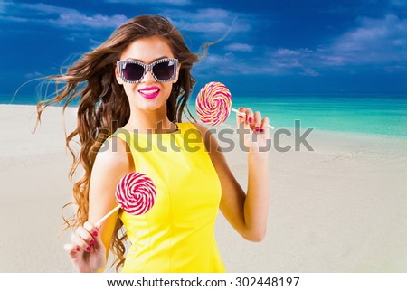 Portrait pretty fun joy sexy smiling brunette woman, has yellow dress, sunglasses, tan face and  body, holding lollipop. Sun tropical hot blue sea water resort. Travel rest nature. Amazing lifestyle.