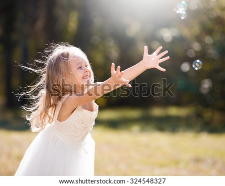 Beautiful little blonde girl, has happy fun cheerful smiling face, white dress, soap bubble blower. Portrait nature.