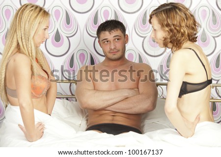 man sitting between two frustrated women in bed - focus on man