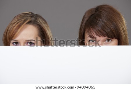 two women peeking over edge of blank empty paper billboard with copy space for text