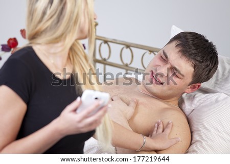 Sick man with ache on bed, worried woman holding phone at his side.