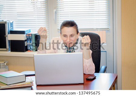 A fifty years old woman working in office with a laptop is very angry with modern technologies. She shows closed fists because she is very upset.