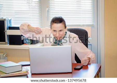 A fifty years old and emotional woman working in office with a laptop is very confused with the modern technology. She shows the fists because she is very angry.