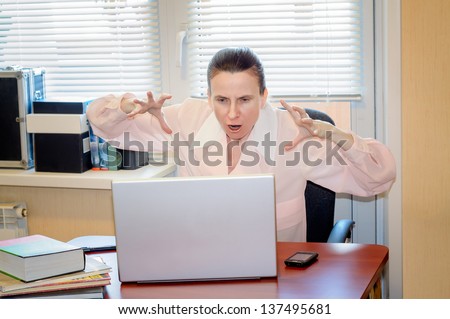 A fifty years old and emotional woman working in office with a laptop is very stressed with the modern technology. She shows the hands like claws because she is very angry.
