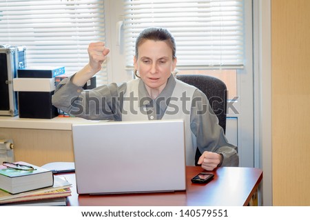 A fifty years old and emotional woman working in office with a laptop is very frustrated with the modern technology. She beats the fist on the table because she is very angry.