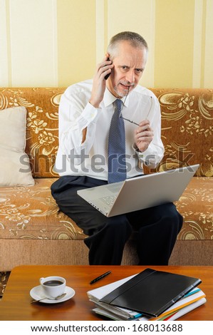 A handsome worried senior businessman working with computer at home, calling to a customer with his mobile phone and ready to drink a hot cup of black coffee