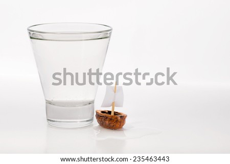 Closeup of a walnut shell boat with a sail, close to a transparent glass full of water or alcohol.