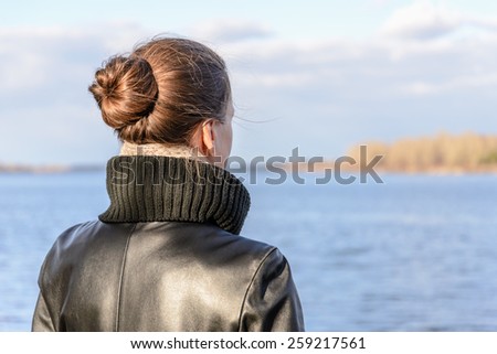 A woman with a chignon and a black leather coat is watching the landscape close to the lake or the river during a sunny day with big white clouds