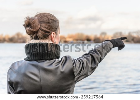 An adult with a chignon and wearing a black leather coat stays close to the river and point her finger to indicate something interesting on the opposite bank