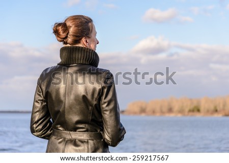 A woman with a chignon and a black leather coat is watching the landscape close to the lake or the river during a sunny day with big white clouds