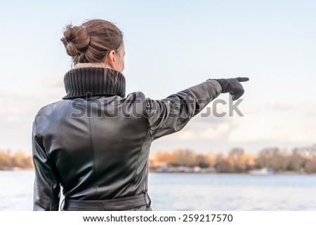 An adult with a chignon and wearing a black leather coat stays close to the river and point her finger to indicate something interesting on the opposite bank