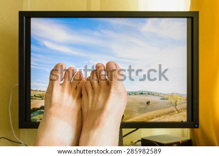 Man\'s feet in front of a computer monitor with a nice sunny italian landscape with a cloudy sky to symbolize the wish of holidays rest