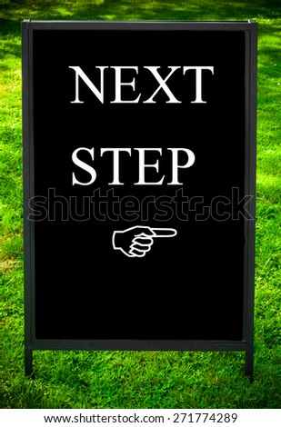 NEXT STEP message and hand pointing to the right on sidewalk blackboard sign against green grass background. Copy Space available. Concept image