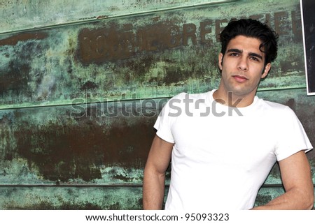 Male model in white shirt leaning against rusty green wall.
