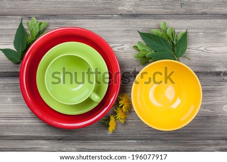 Top view empty red plate, yellow bowl and green cup on rustic wooden table with spring leaf and flower