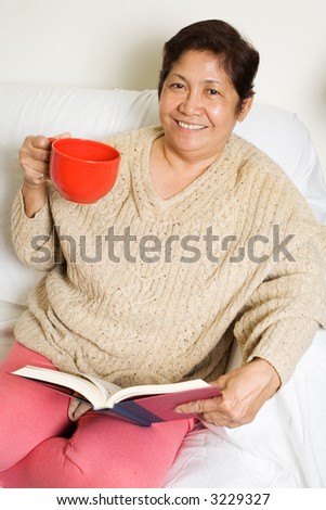 Senior asian woman relaxing at home drinking a cup of coffee while reading a book smiling while looking at the camera