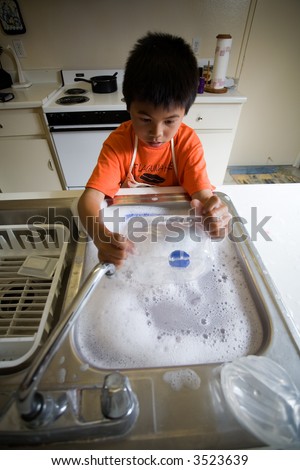 Young independent asian boy wearing an apron standing in front of kitchen sink full of soapy water washing the dishes