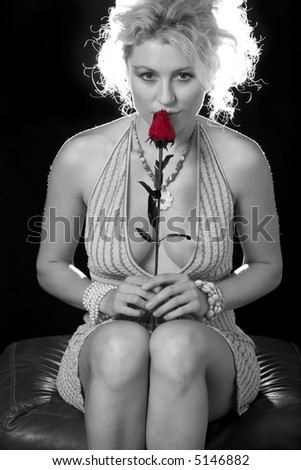 Full body of an attractive woman with curly blond hair in a sexy low cut dress sitting over black holding a red rose