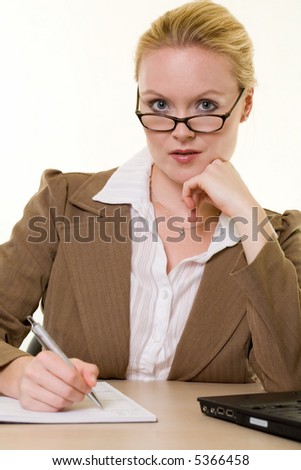 Attractive blond woman dressed in professional business attire wearing dark framed eyeglasses sitting at a desk writing in an agenda resting chin on hand