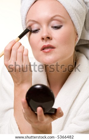 Attractive blond woman applying makeup wearing white robe and towel over hair