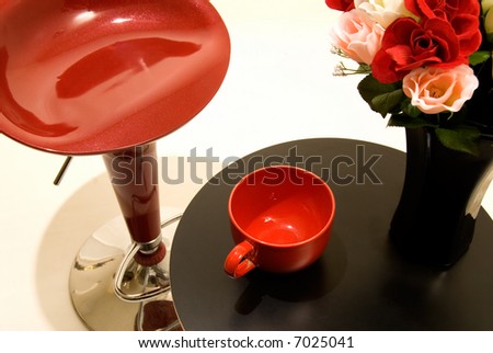Small end table with a black vase full of red and pink roses beside a big red coffee mug and unique red bar stool
