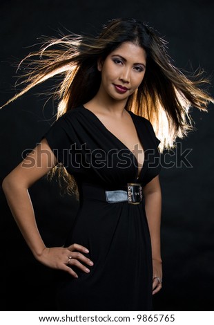 Close up of face of an attractive long haired brunette Hawaiian woman wearing a black low cut top with hair back lit and blowing over black