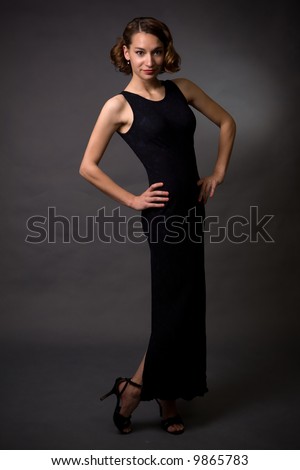 Full body of young pretty brunette woman modeling long black dress while standing on grey background