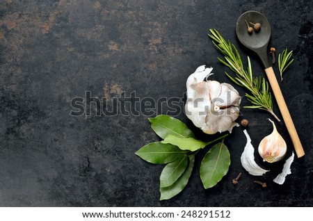 Dark culinary background with a wooden spoon along with garlic, rosemary, bay leaves,  pepper and some cloves pictured on it