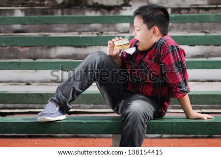 asian boy eating muffin sandwich on a green concrete bench
