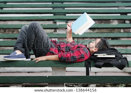 an asian boy lying on a green concrete bench, reading a book