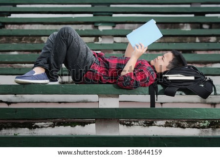 an asian boy lying on a green concrete bench, reading a book