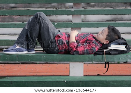 an asian boy sleeping on a green concrete bench with backpack under his head
